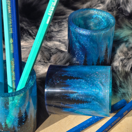 Handgefertigter Stiftehalter aus dunkelblauem Resin mit glitzernden Polarlichtern und schwarzer Tannen-Silhouette, gefüllt mit blauen Buntstiften.