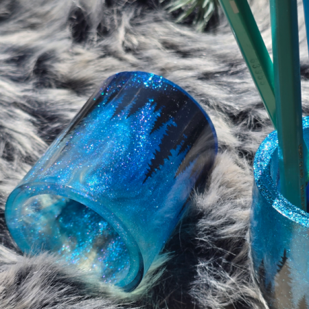 Handgefertigter Stiftehalter aus dunkelblauem Resin mit glitzernden Polarlichtern und schwarzer Tannen-Silhouette, gefüllt mit blauen Buntstiften.